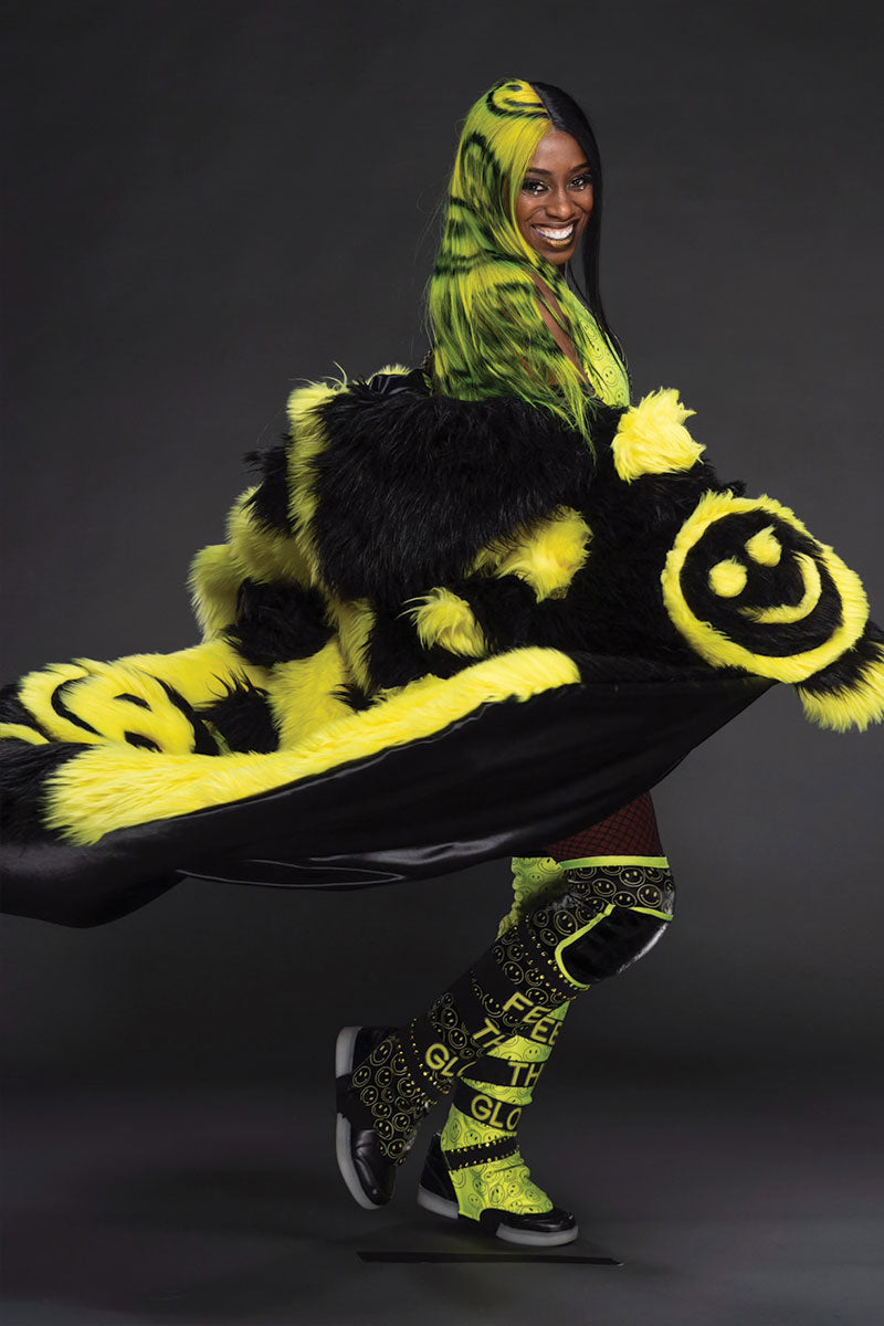 Person wearing a neon yellow and black outfit with smiley faces. They are also wearing a half black half neon yellow wig with smiley faces painted into the hair.