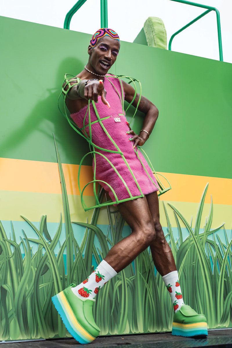 Model wearing a pink dress and colorful shoes with a green and yellow striped background. The model is wearing a patterened buzzed wig in vibrant colors.