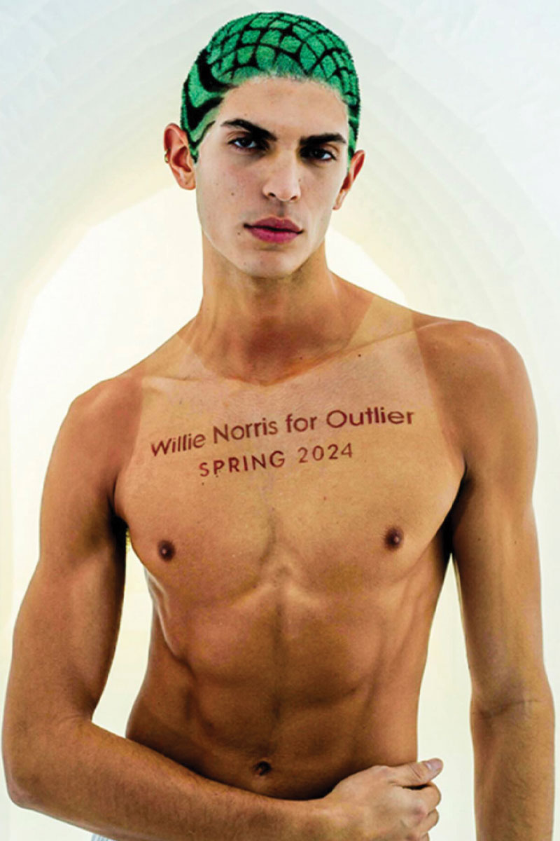 a model wearing a green and black buzzed wig. they are shirtless and their chest says willie noris for outlier spring 2024