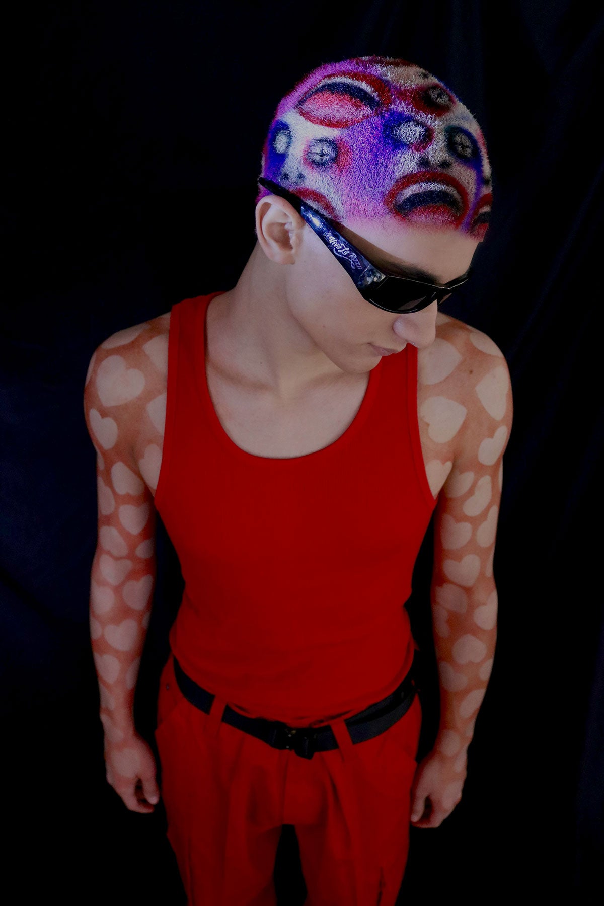 a model in a red outfit wearing a buzzed, colorful wig with faces painted into it
