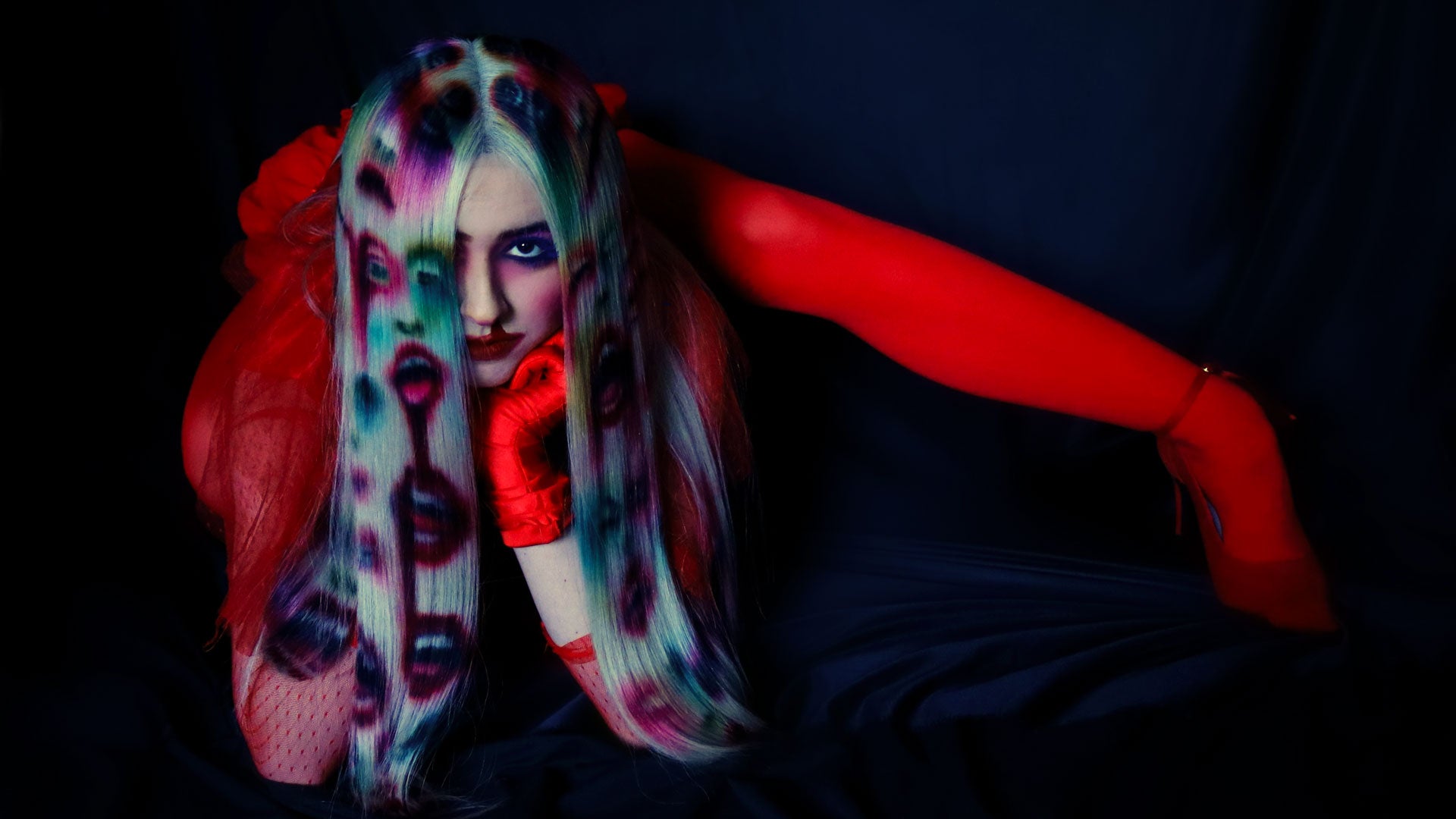 An image of a model in red clothing wearing a white blonde wig. Faces and mouths are painted into the wig