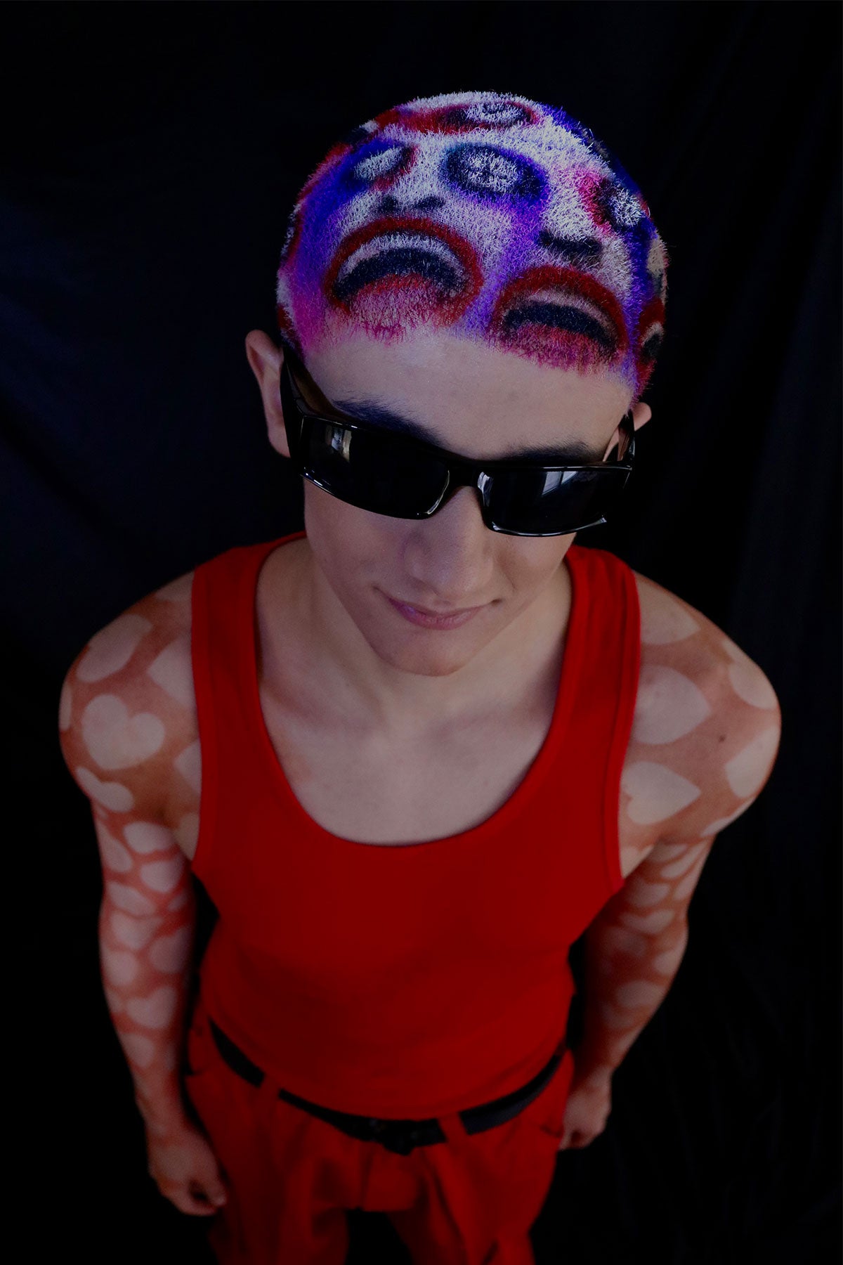 a model in a red outfit wearing a buzzed, colorful wig with faces painted into it