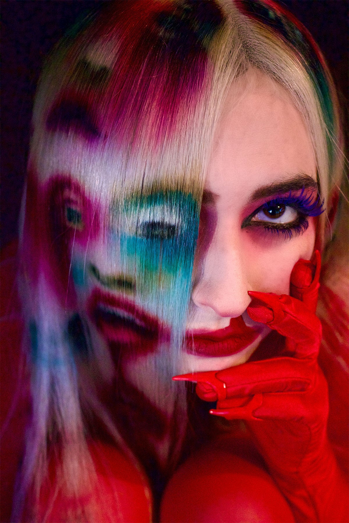 A close up of a model in red clothing wearing a white blonde wig. Faces and mouths are painted into the wig