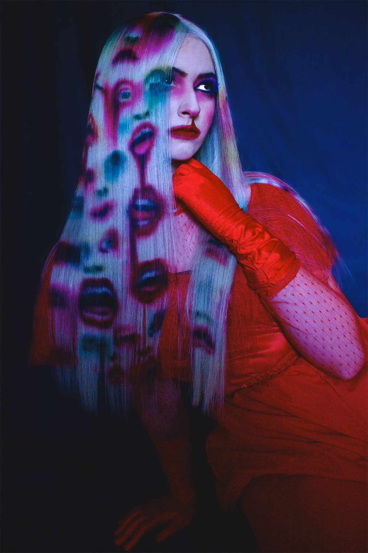 A front side view of a model in red clothing wearing a white blonde wig. Faces and mouths are painted into the wig