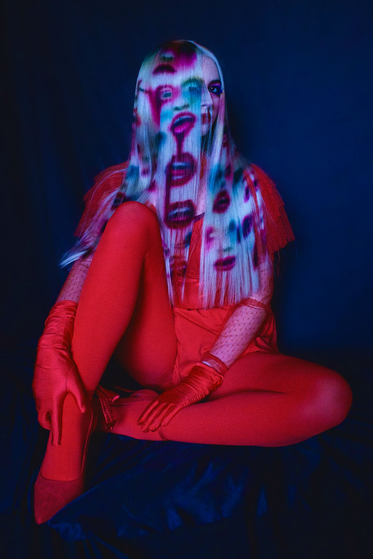 A front side view of a model in red clothing wearing a white blonde wig. Faces and mouths are painted into the wig