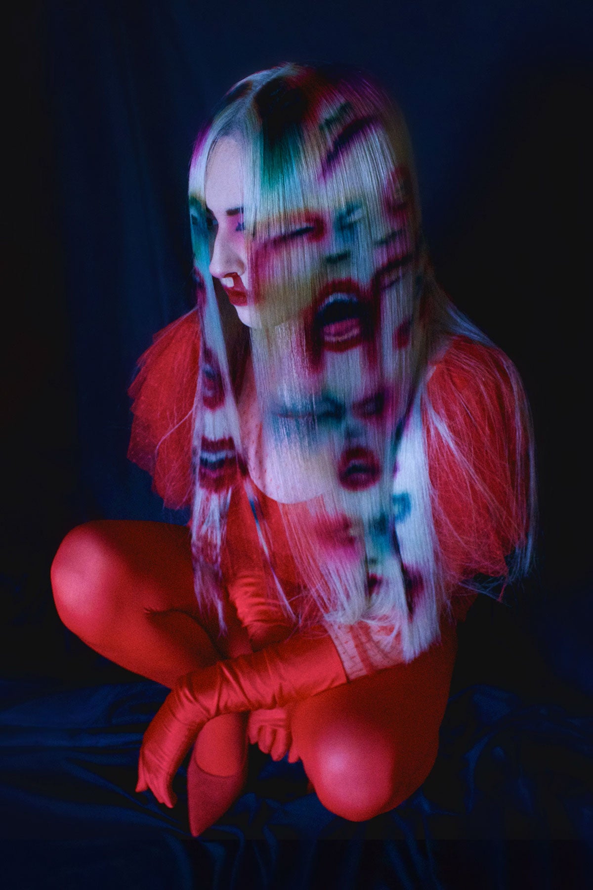 A front side view of a model in red clothing wearing a white blonde wig. Faces and mouths are painted into the wig