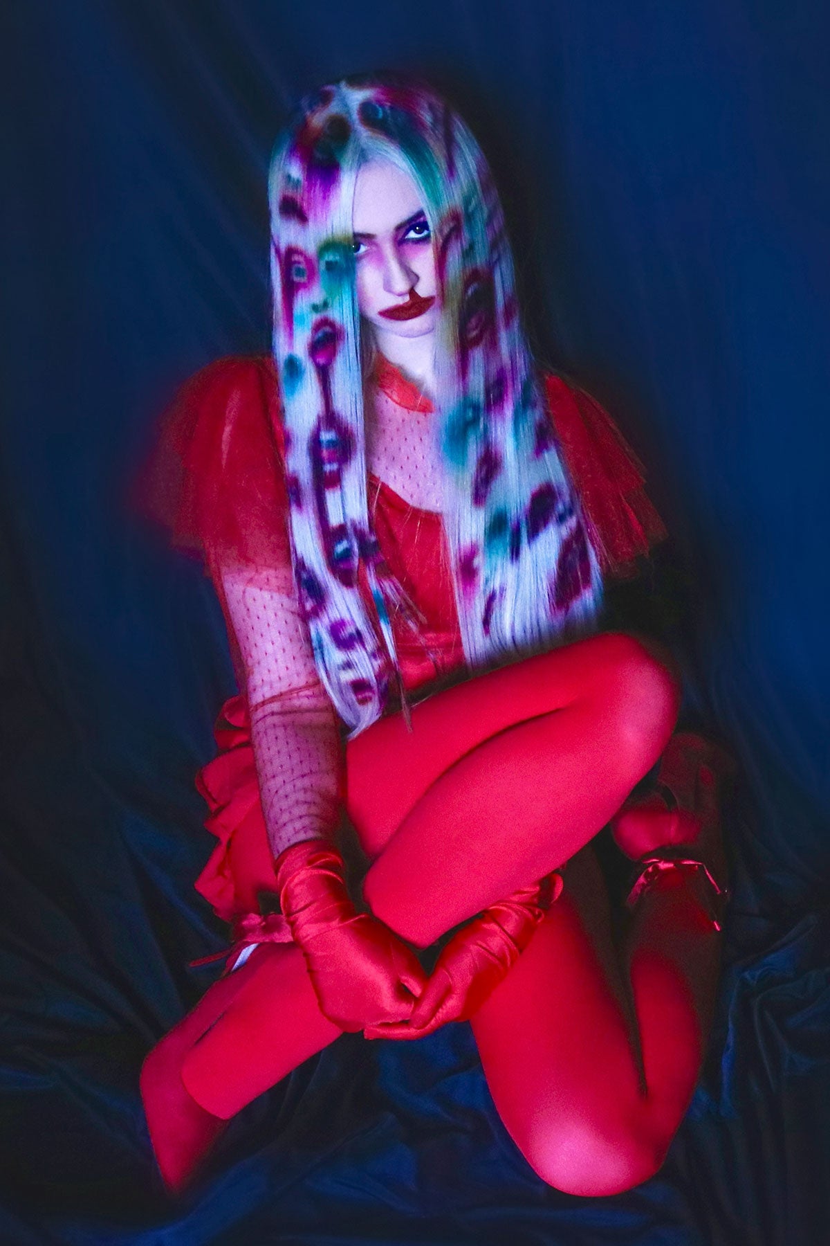 A front view of a model in red clothing wearing a white blonde wig. Faces and mouths are painted into the wig