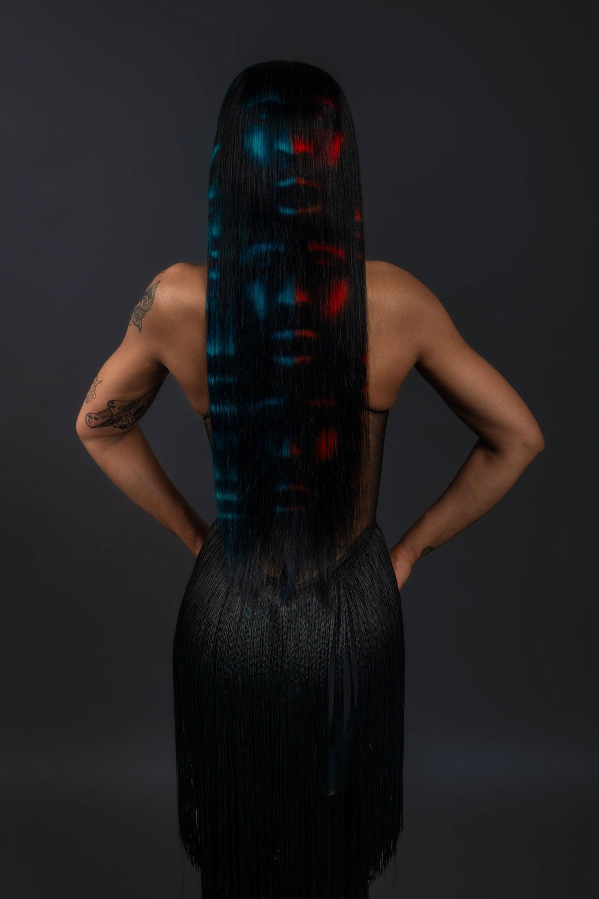 a back view of a model wearing a black outfit and a black waist length wig with blue and red faces painted into it