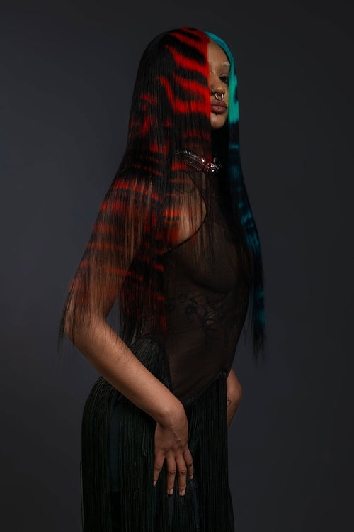 a side view of a model wearing a black outfit and a black waist length wig with blue and red faces painted into it