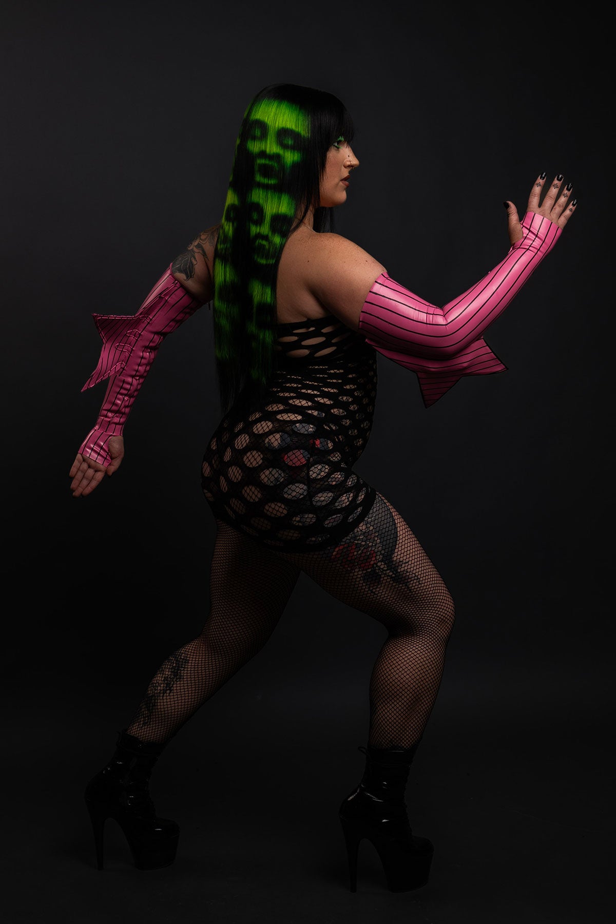 a side view of a model wearing a black and green zombie wig and a fishnet dress with black boots