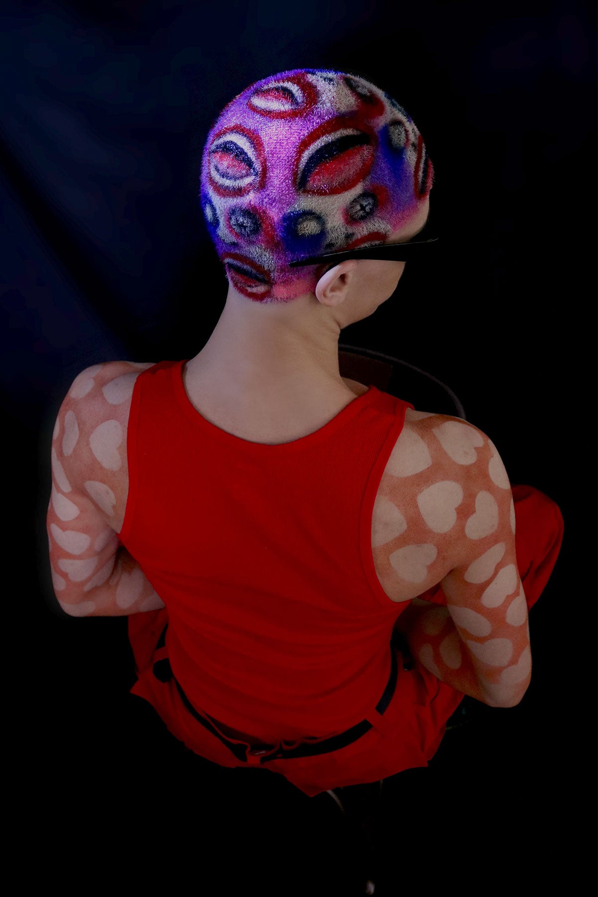 a model in a red outfit wearing a buzzed, colorful wig with faces painted into it