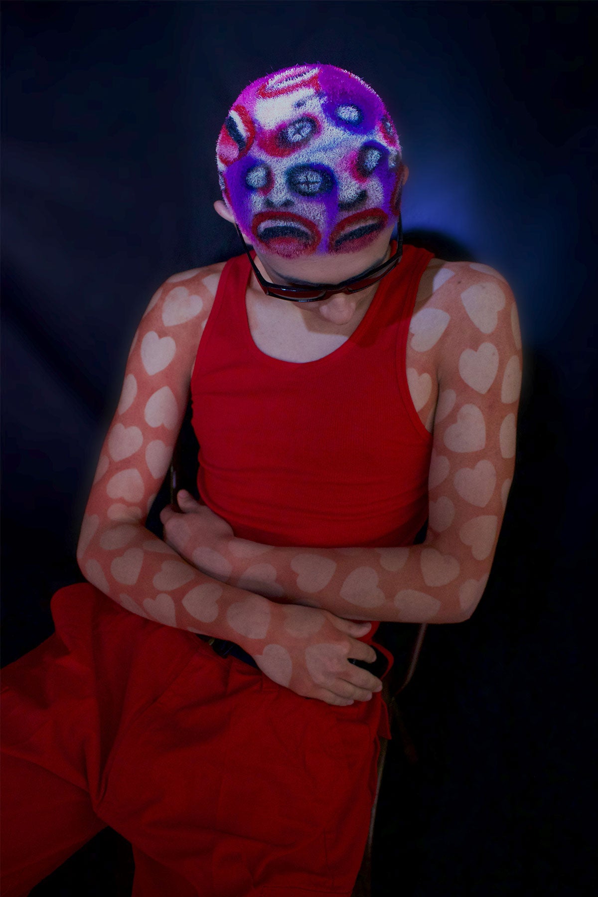 a model in a red outfit wearing a buzzed, colorful wig with faces painted into it