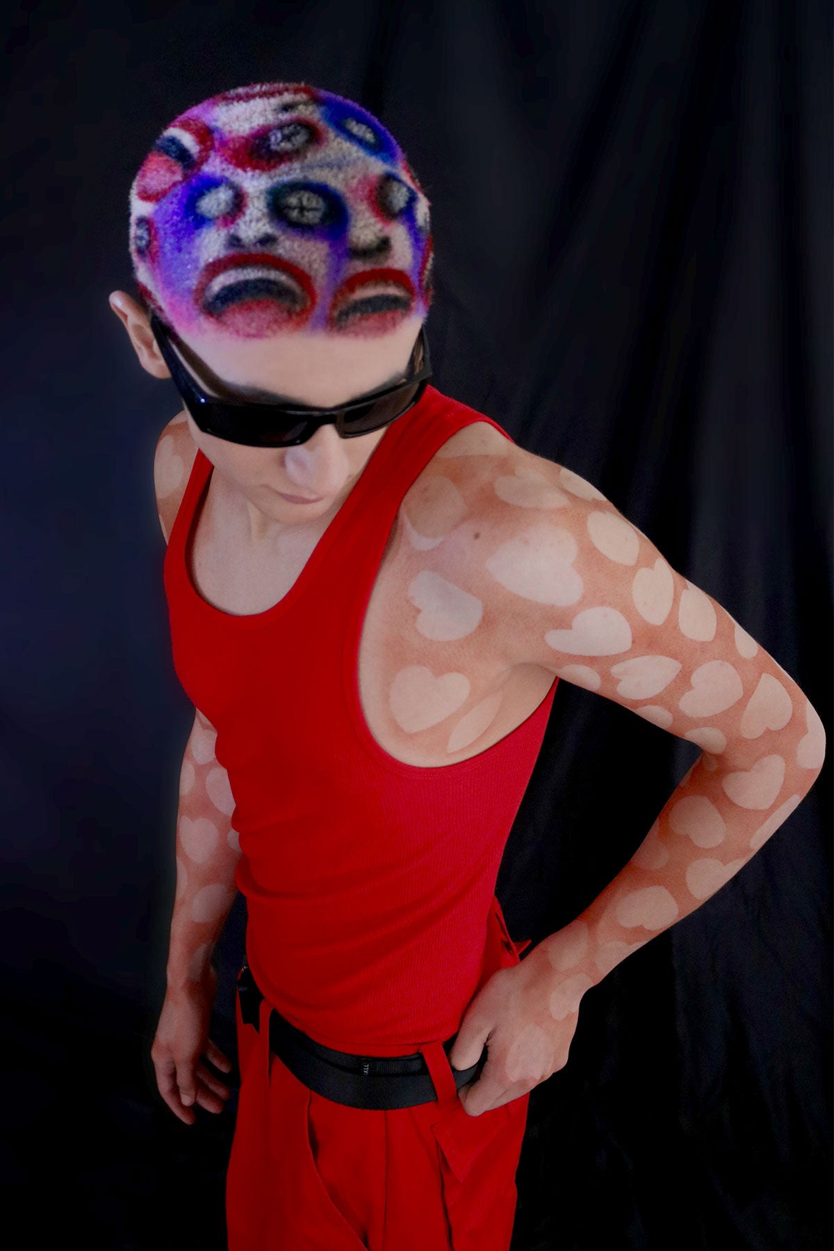 a model in a red outfit wearing a buzzed, colorful wig with faces painted into it