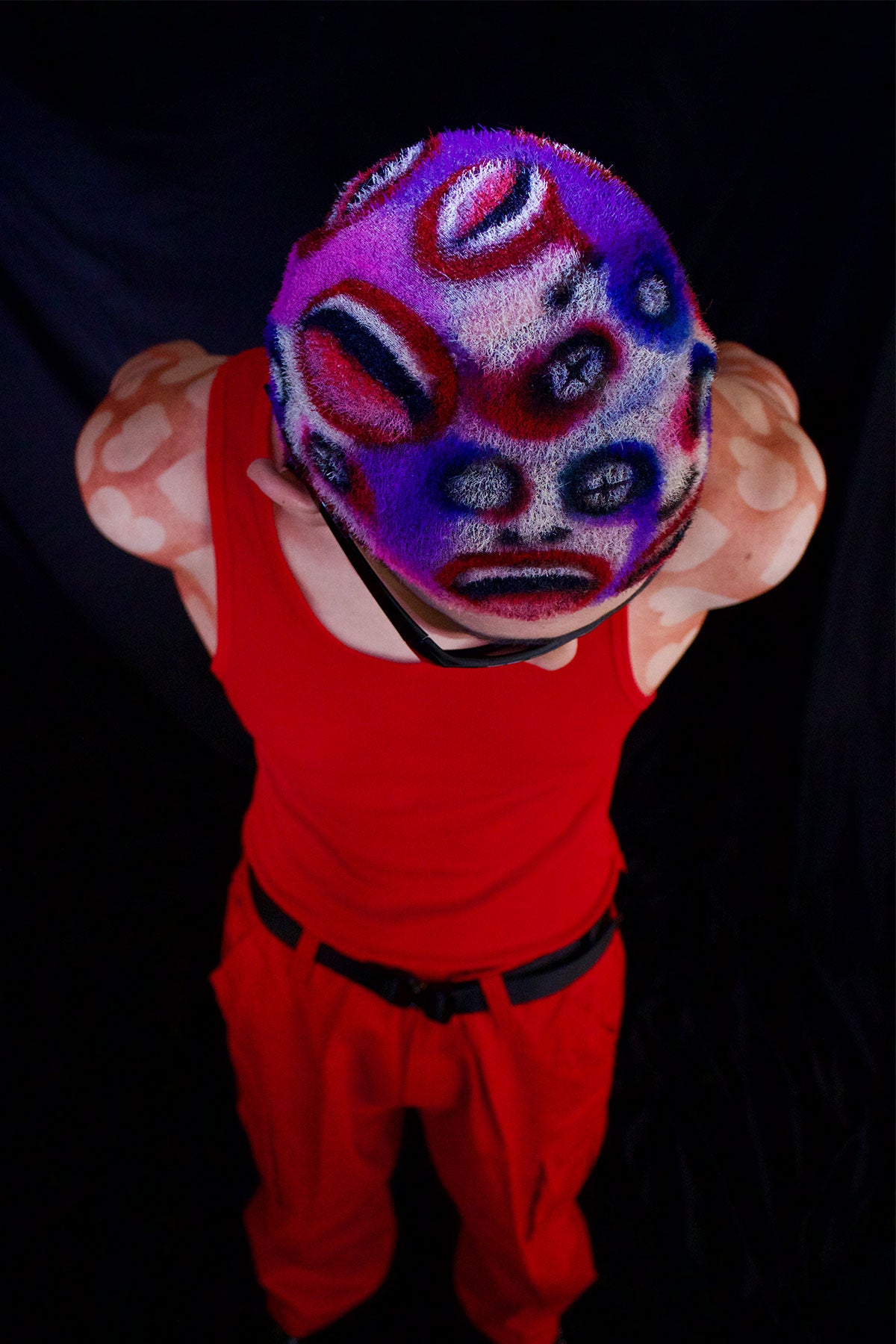 a model in a red outfit wearing a buzzed, colorful wig with faces painted into it