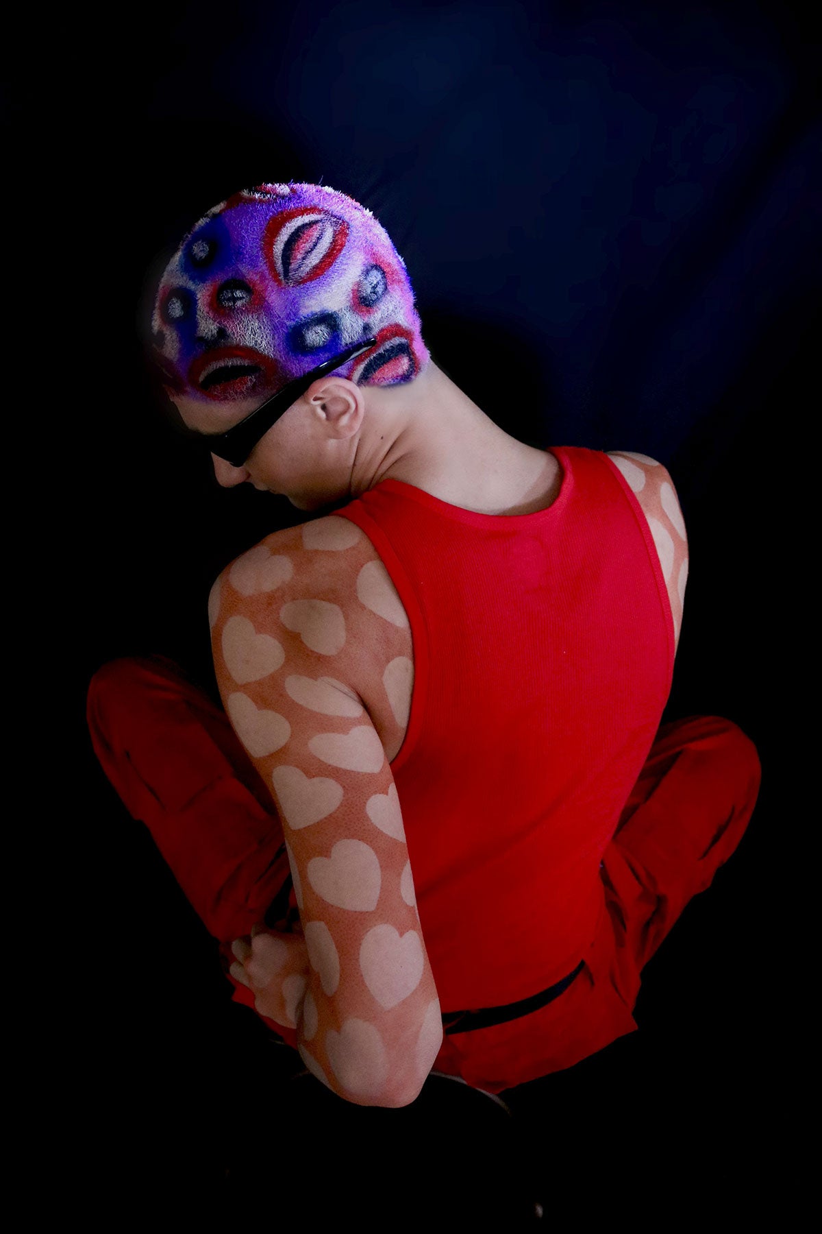 a model in a red outfit wearing a buzzed, colorful wig with faces painted into it