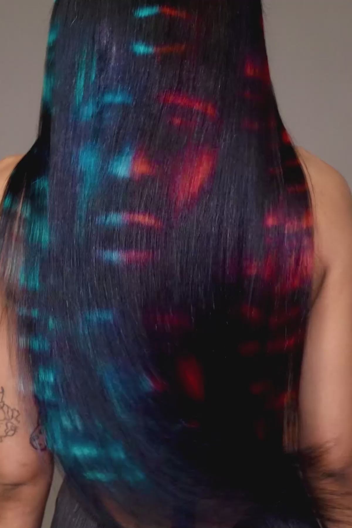 a back view of a model wearing a black outfit and a black waist length wig with blue and red faces painted into it, shaking her head and making the wig move