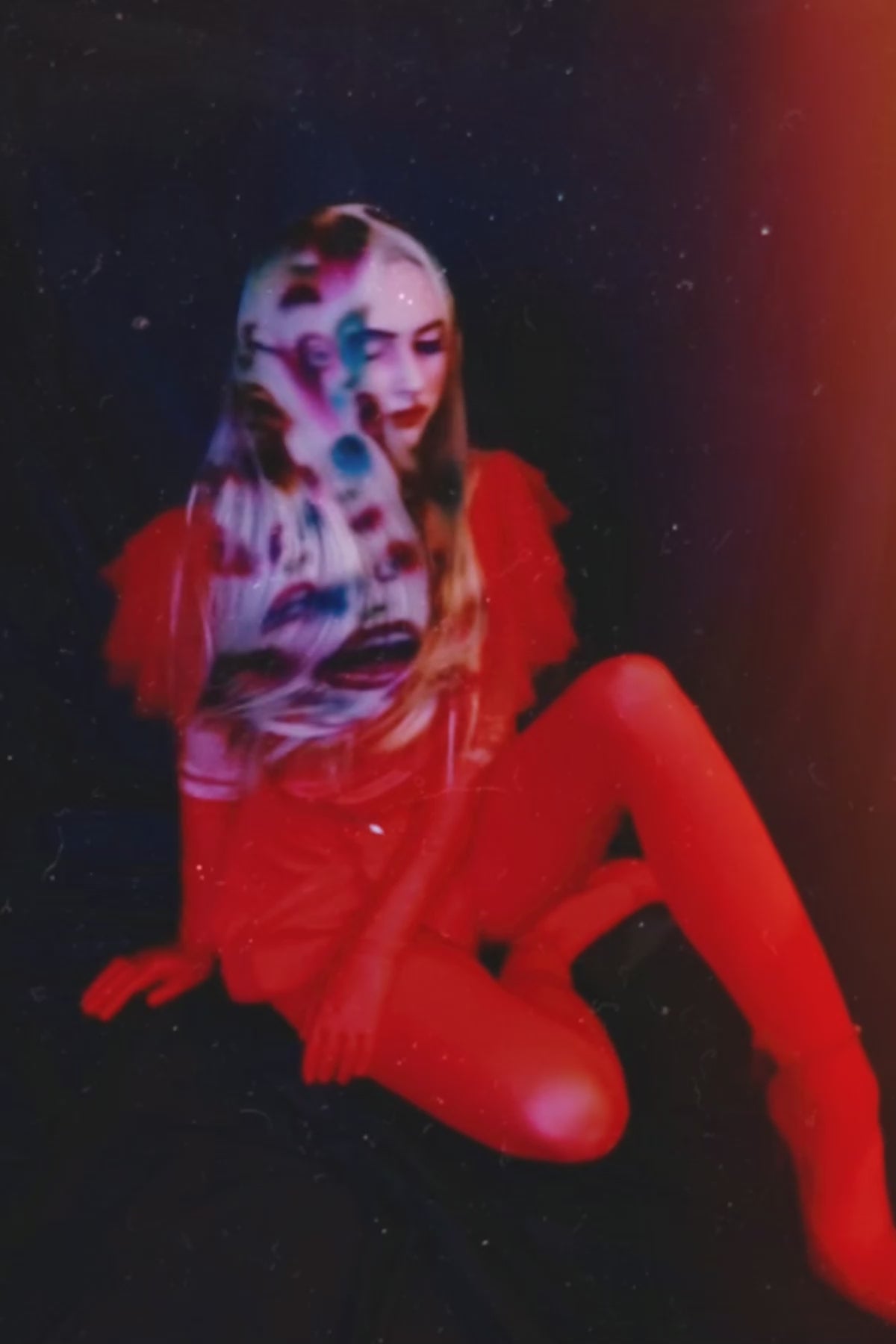 A model in a red shirt and white blonde wig with faces and mouths painted into the wig. The model is moving her head back and forth so the wig moves