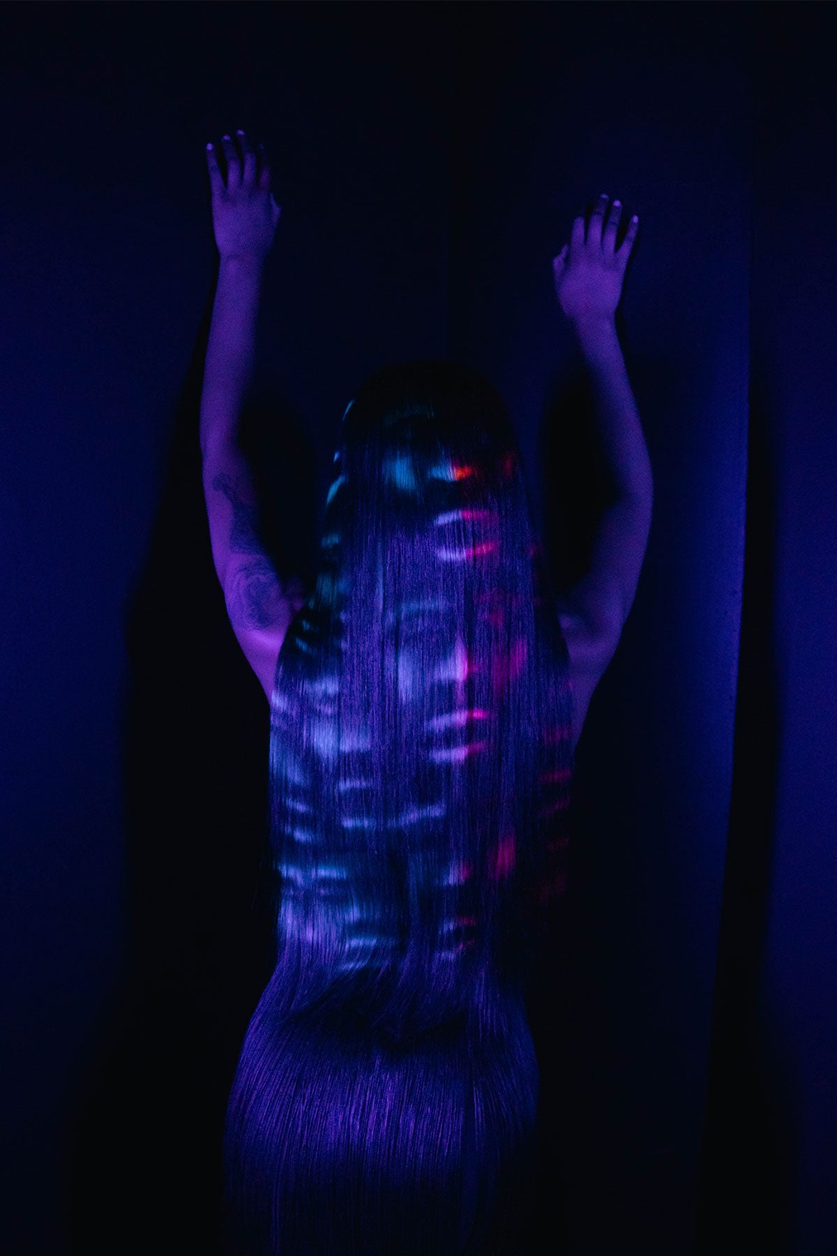 a back view of a model wearing a black outfit and a black waist length wig with blue and red faces painted into it, under black light